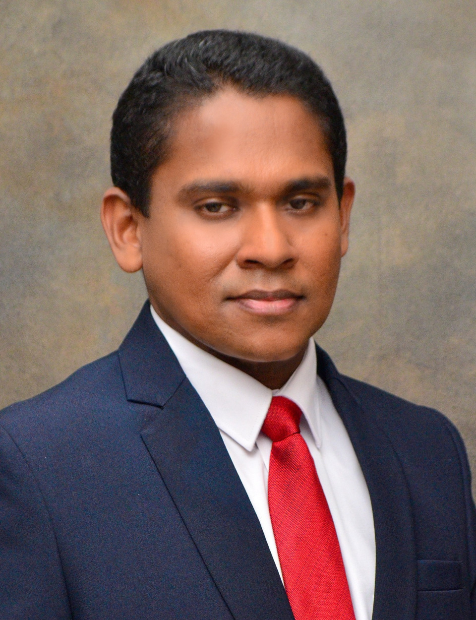 Professional, friendly portrait of a South Asian male consultant psychiatrist in a clinic office. Neutral light background, soft natural lighting, white coat and stethoscope, warm approachable smile, photographic realism.